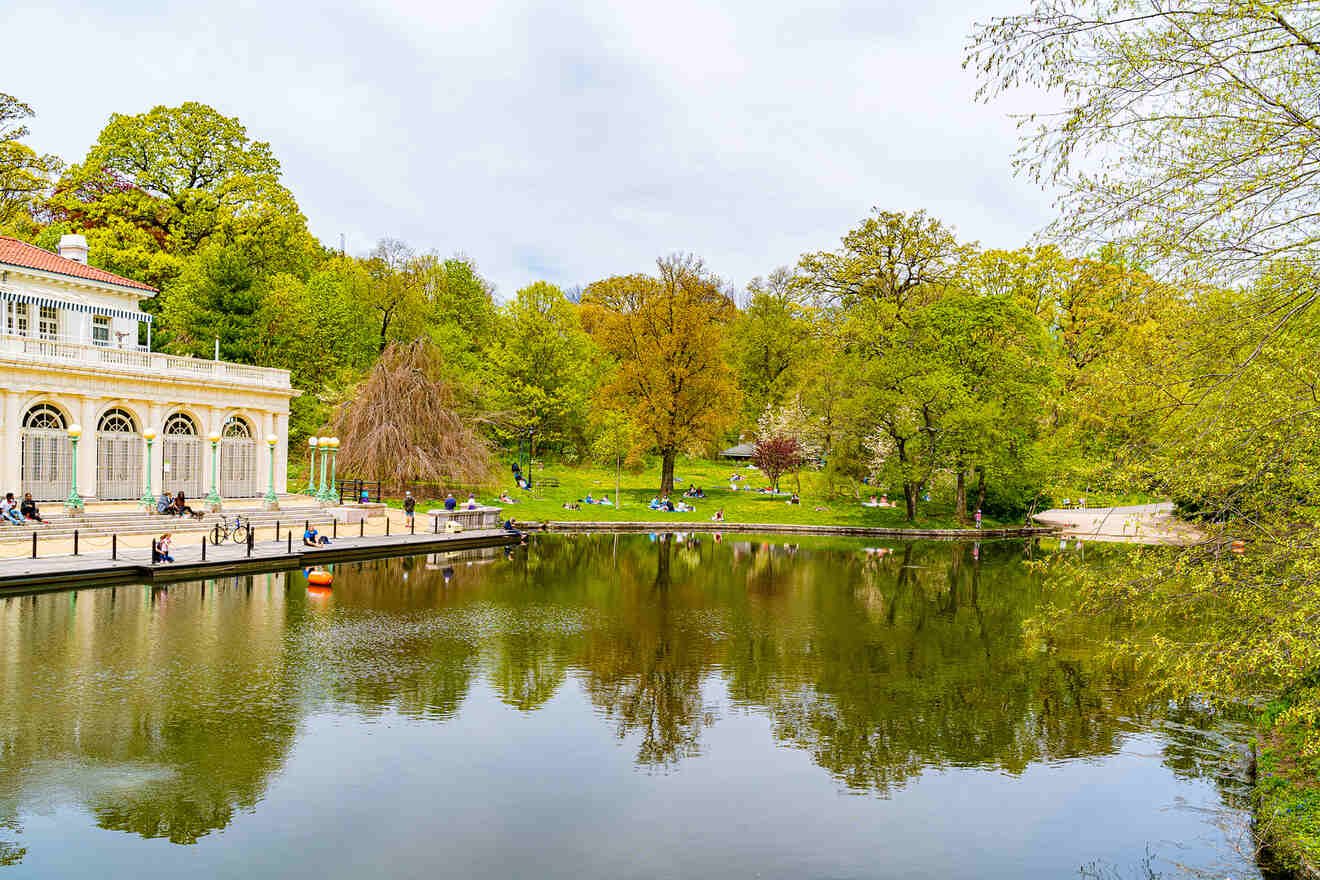 A serene park scene with people relaxing by a pond. Trees are reflected in the water, and a classical building stands to the left.